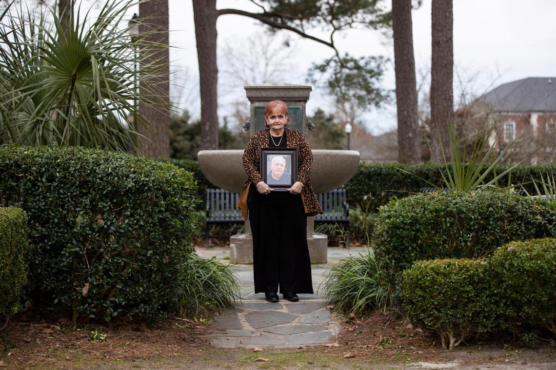 Karen Lovette poses for a portrait holding a photograph of her husband Al Lovette in Camden, South Carolina on Tuesday, March 2, 2021. Before her husband died from the coronavirus, the Lovettes would sit by this fountain at Hampton Park after lunch.