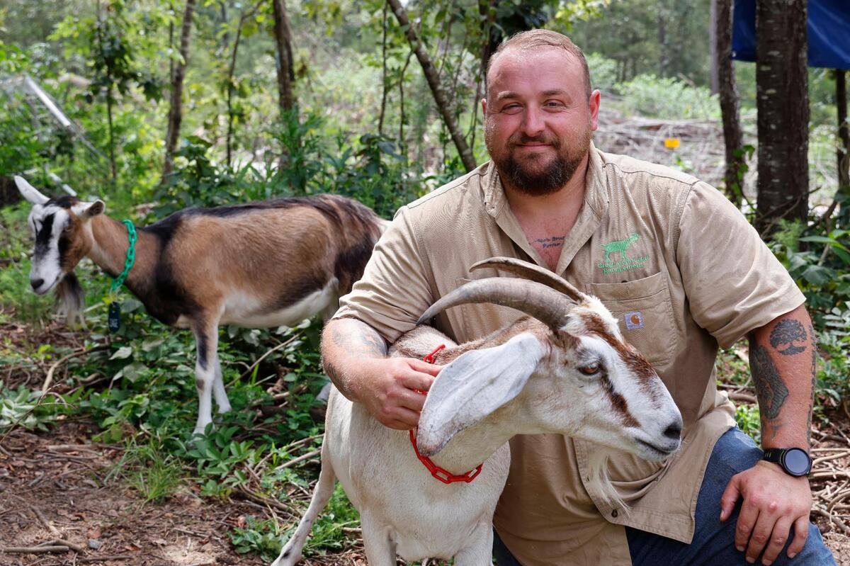 Jacob Porter, owner of goatscaping company Green Goat Land Management, poses with his goats at a home in Gadsden, South Carolina on Thursday, July 27, 2023. His business brings goats, who will eat overgrowth over several days.