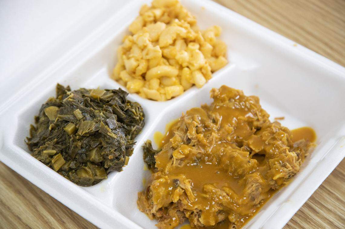 A close-up of a classic Southern barbecue plate in a white styrofoam container. The main compartment is filled with pulled pork covered in a thick, mustard-colored sauce. The two side compartments hold a scoop of creamy macaroni and cheese and a portion of dark, cooked collard greens.