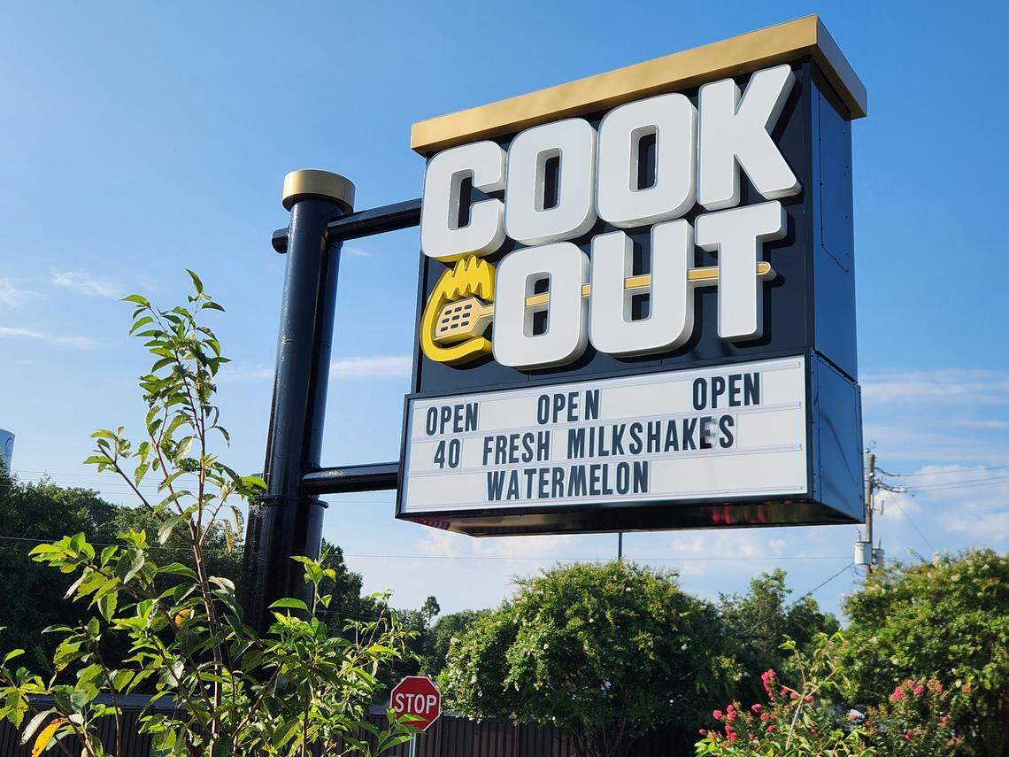 A photograph of a Cook Out restaurant sign against a blue sky with some green foliage in the foreground. The sign has large, backlit letters spelling “COOK OUT” at the top. Below, a lit-up message board reads “OPEN,” “OPEN,” “OPEN,” “40 FRESH MILKSHAKES,” and “WATERMELON.” A red stop sign is partially visible at the bottom of the frame.