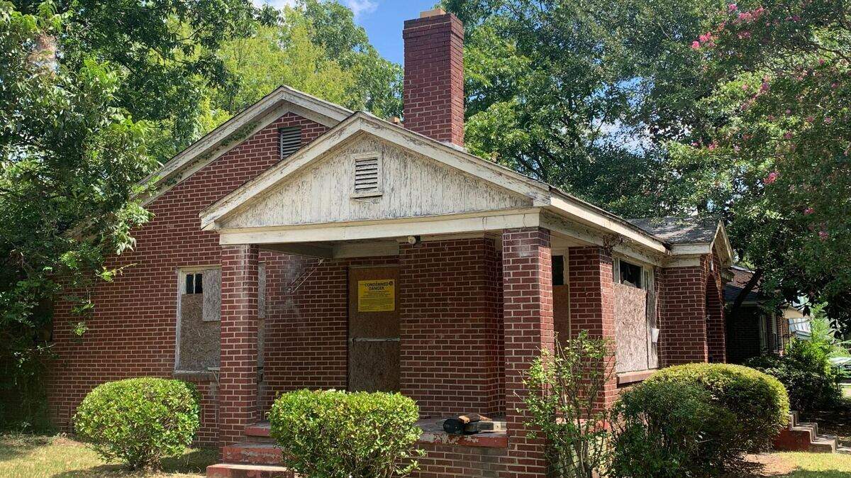 A photo of a red brick house and its side yard. The house is slightly tarnished and has a large yellow poster attached saying "CONDEMNED. DANGER."