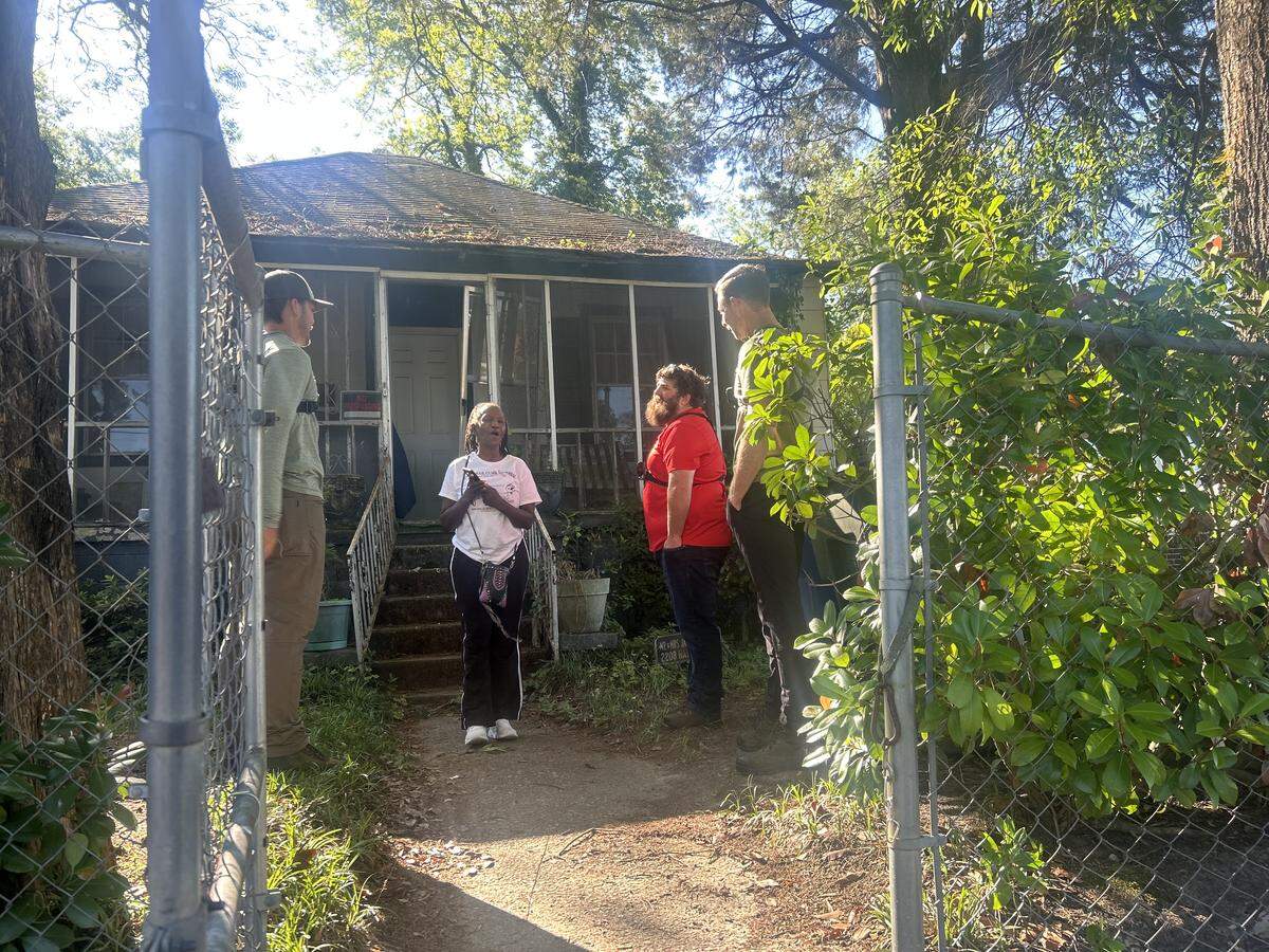 Three men stand around a surprised woman in front of her house.