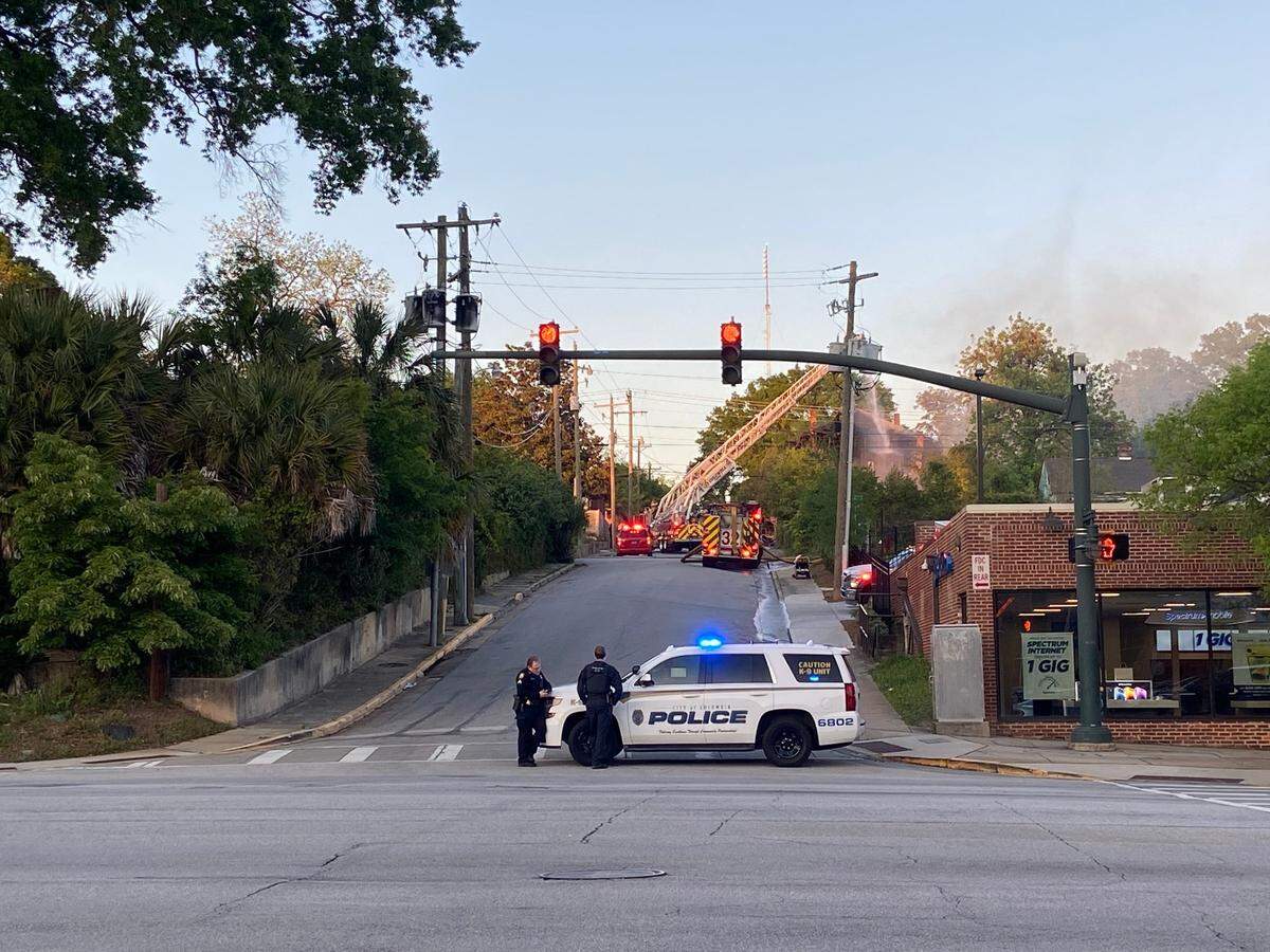 Smoke rises from a burning house and fire engines occupy the street outside. 
