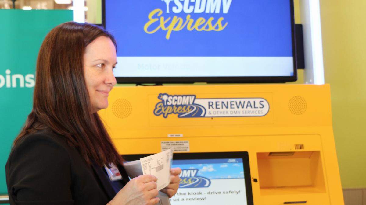 A woman with reddish-brown hair standing in front of a large yellow-orange kiosk with a virtual display. The woman is holding up a piece of paper -- a vehicle registration document printed by the kiosk. 