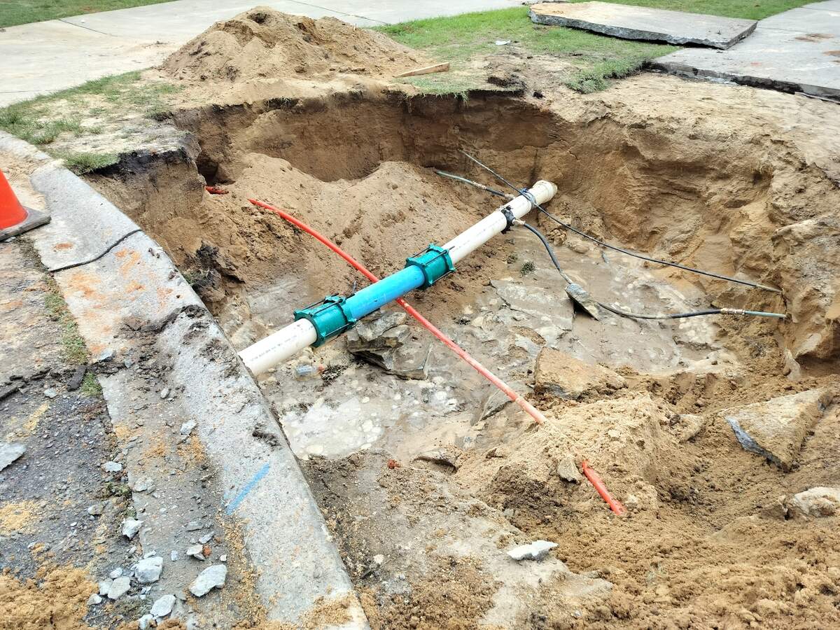 A photo showing a wide sinkhole where someone's driveway once was. There is an exposed pipe in the middle of the sandy hole. 