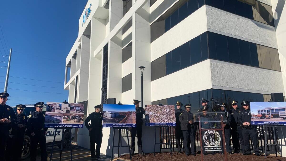 A group of police officers stand outside of an empty white office building with an old Aflac logo on the wall. The police officers are standing in a line next to renderings of what the new facility will look like. 