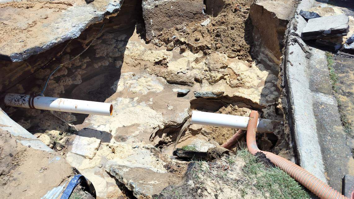 A photo showing a wide sinkhole where someone's driveway once was. There is an exposed pipe in the middle of the sandy hole. 
