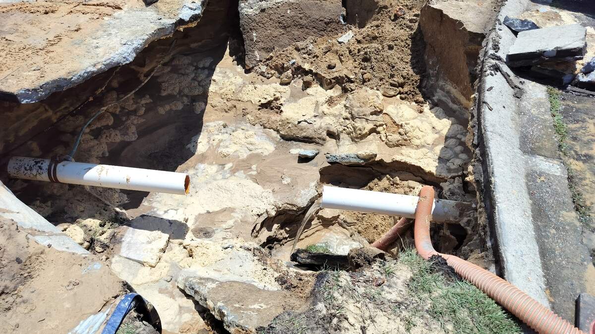 A photo showing a wide sinkhole where someone's driveway once was. There is an exposed pipe in the middle of the sandy hole. 