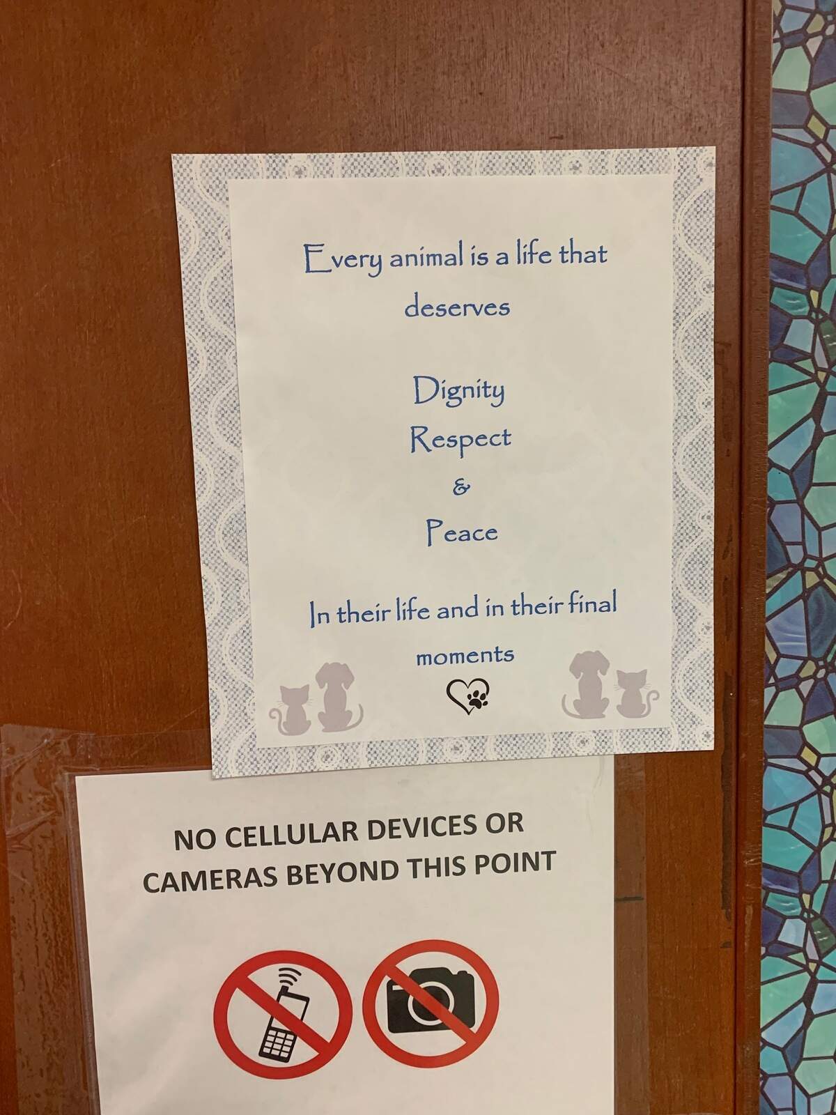 Two posters are attached to a wooden door. One reads "Every animal is a life that deserves dignity respect and peace in their life and in their final moments." The other reads "no cellular devices or cameras beyond this point." 