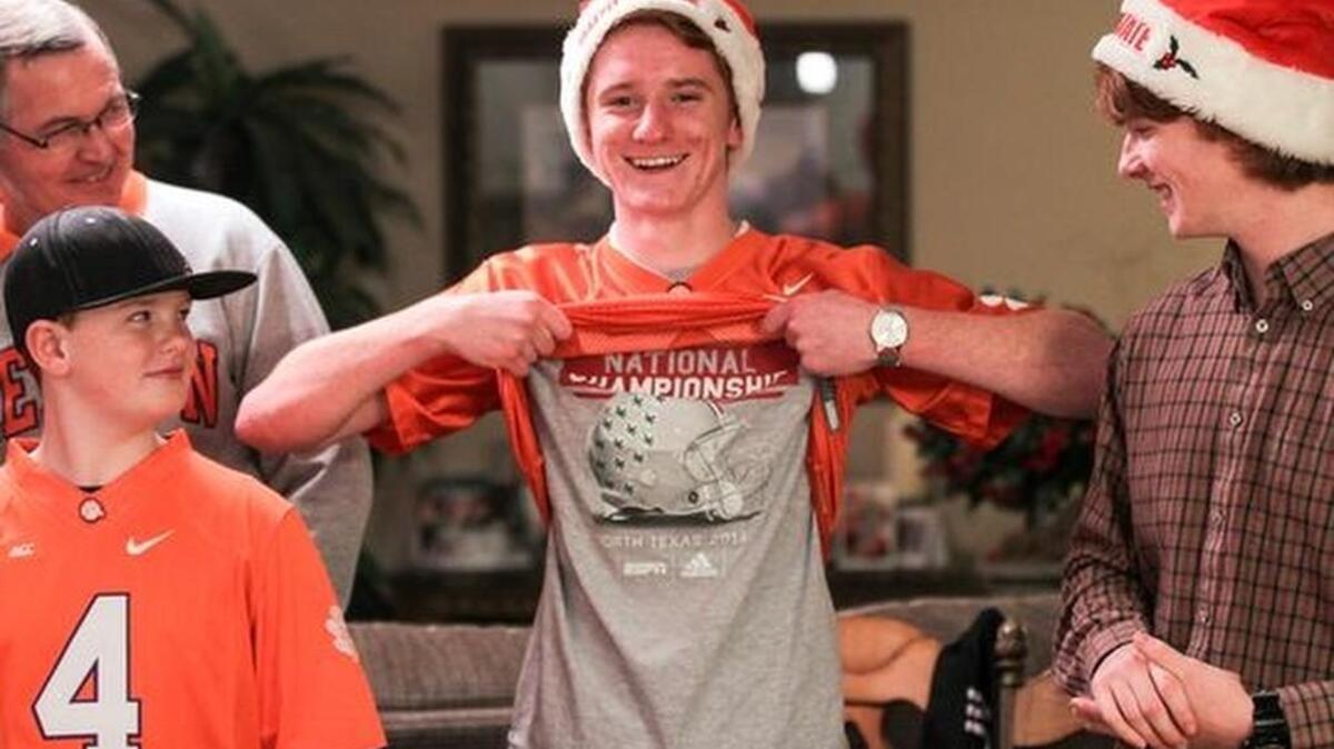 Bob King, left, watches Brent Merlo show off an Ohio State 2013 National Championship T-shirt underneath a Clemson Tiger’s jersey. Brent, the ‘people pleaser’ in the family is flanked by brothers Derrick, left, and Chris, right, whose families are split over which team they support.