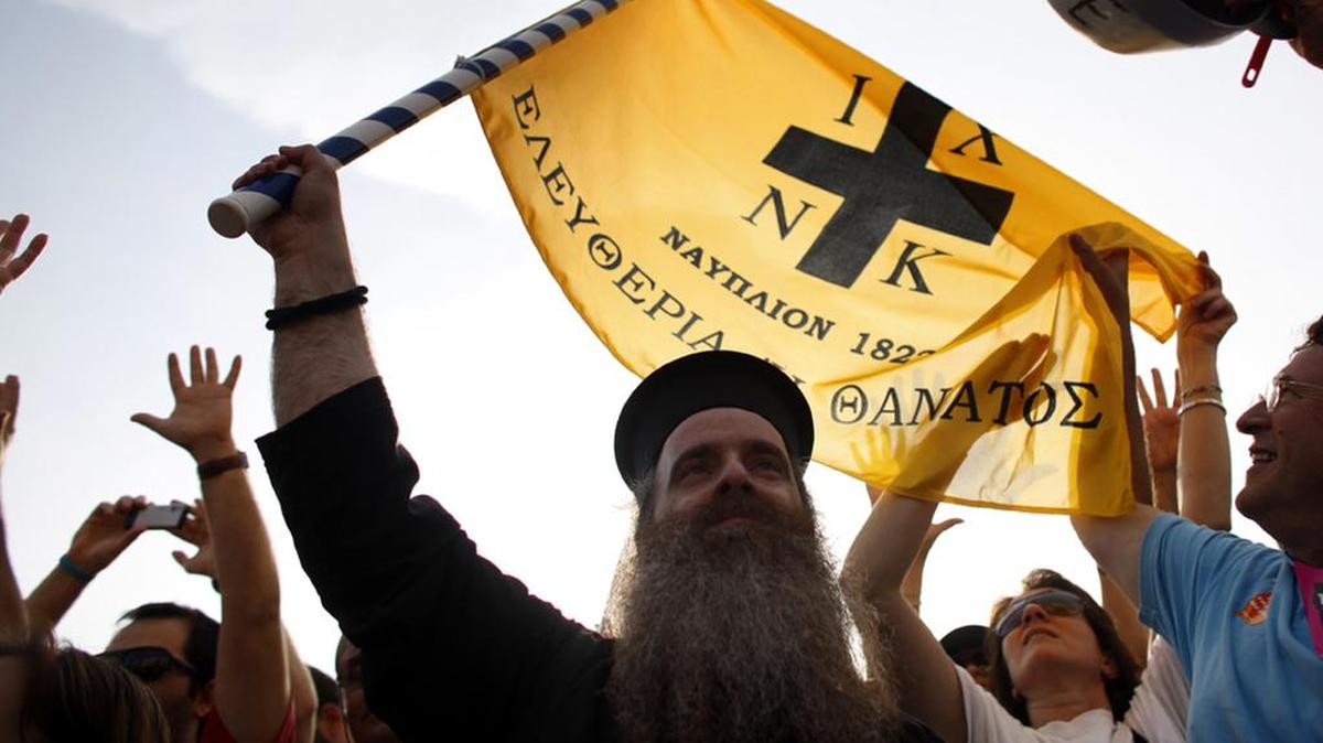 A Greek orthodox priest holds an old flag of the Greek revolution that reads “freedom or death.”